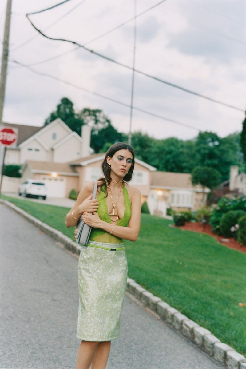 An adult female with long brown hair stands outside wearing a green top and skirt, accessorized with gold jewelry and a handbag.