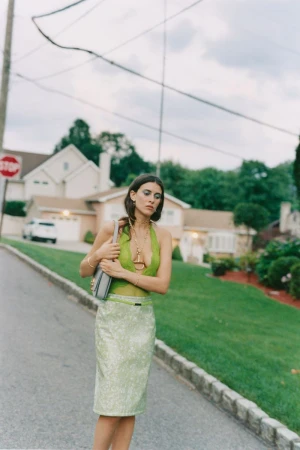 An adult female with long brown hair stands outside wearing a green top and skirt, accessorized with gold jewelry and a handbag.
