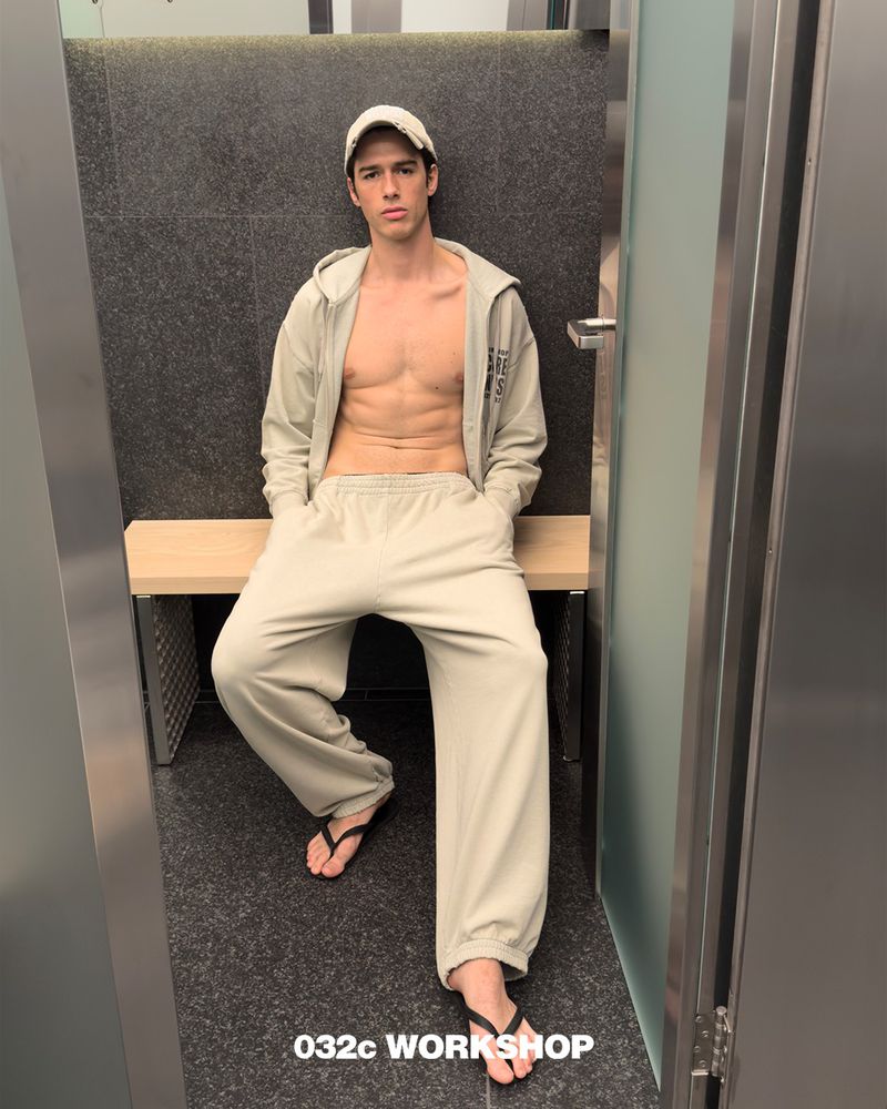 A young adult man in a light-colored sweat suit and baseball cap sits on a bench in an indoor setting, wearing flip-flops. 