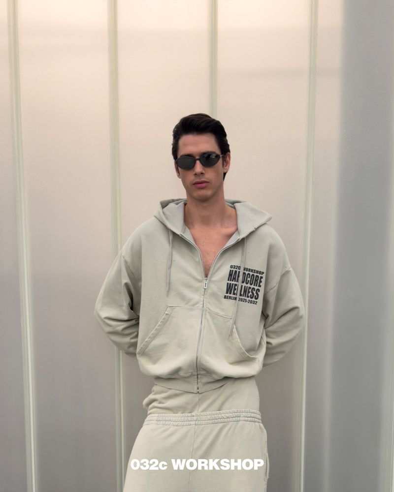 Male model poses in a grey zip hoodie and matching sweatpants against a grey-white paneled wall, wearing oval sunglasses and casual attire.