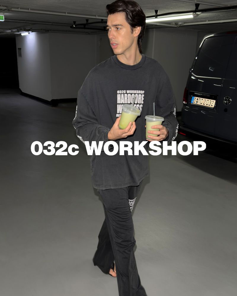 Adult man wearing oversized long-sleeve shirt, black pants, and holding two drinks in an underground parking garage setting.
