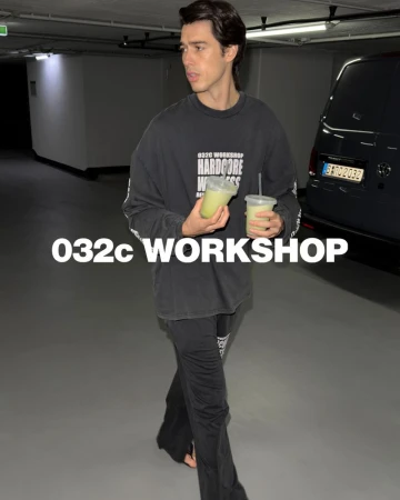 Adult man wearing oversized long-sleeve shirt, black pants, and holding two drinks in an underground parking garage setting.