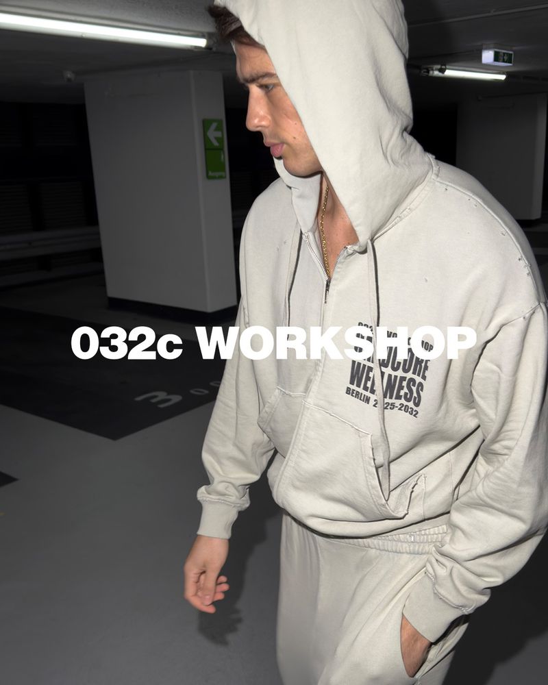 A young adult male in a casual hooded sweatshirt and sweatpants outfit, photographed in an indoor parking garage setting. 