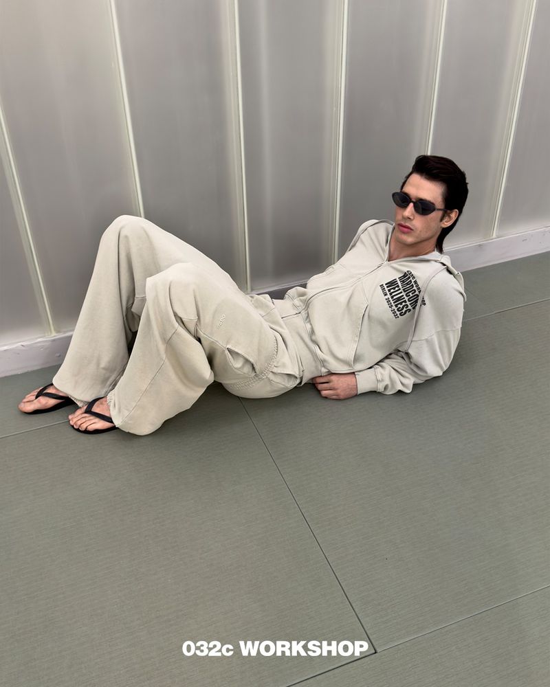 Man in a light-beige hooded sweatshirt and pants poses lying on a grey floor in front of a vertically paneled light wall, wearing sunglasses.