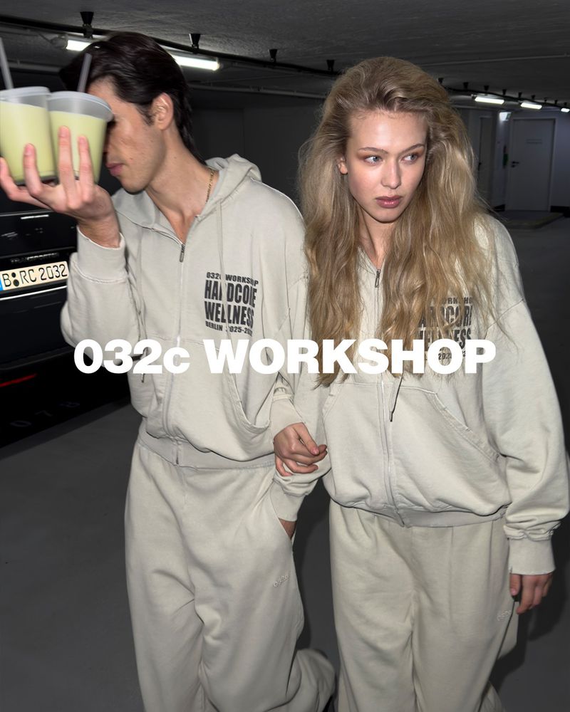 Two fair-skinned adults in matching light tan tracksuits hold smoothie cups in an indoor garage-like setting with muted lighting.