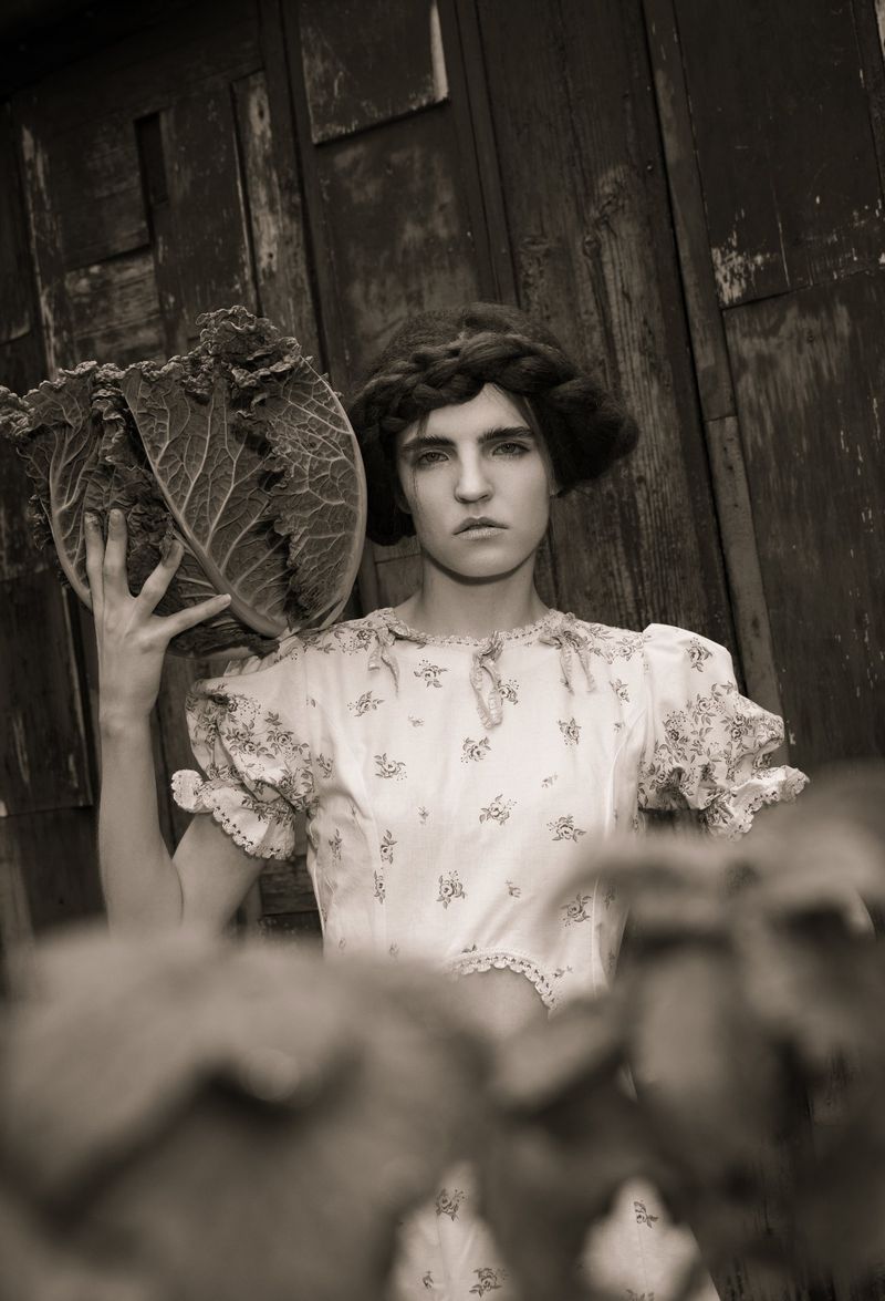 Artistic portrait of a serious woman in a floral dress holding a large cabbage, set against a rustic wooden background.