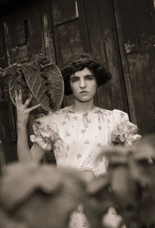 Artistic portrait of a serious woman in a floral dress holding a large cabbage, set against a rustic wooden background.