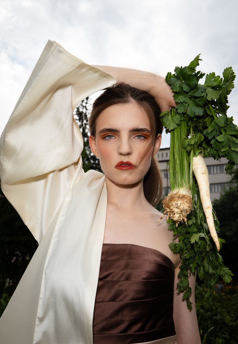 Portrait of a woman holding a large bunch of celery in an outdoor setting, wearing a brown dress and cream-colored robe.