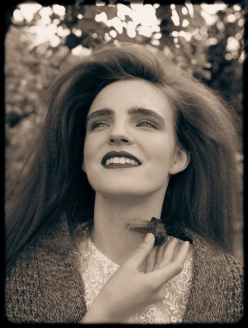 Portrait of a smiling woman with dark hair, wearing a vintage-style dress and cardigan with a beetle on her neck, in a natural setting.