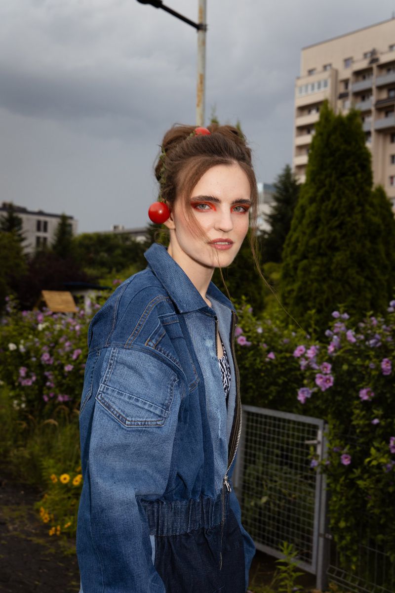 Woman in denim jacket poses in a garden with overcast sky and apartment building background. She has red accessories in her hair.