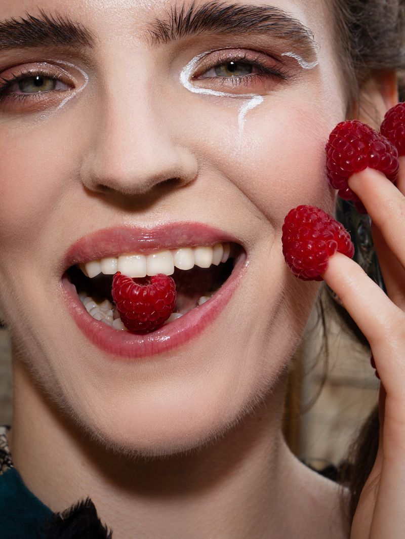 Close-up of a smiling woman with white face makeup holding raspberries near her face and one in her mouth.