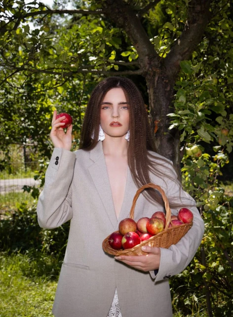 Woman in blazer in orchard holding basket of apples, with apple in hand, wearing white earrings, outdoor, summertime, green foliage backdrop.