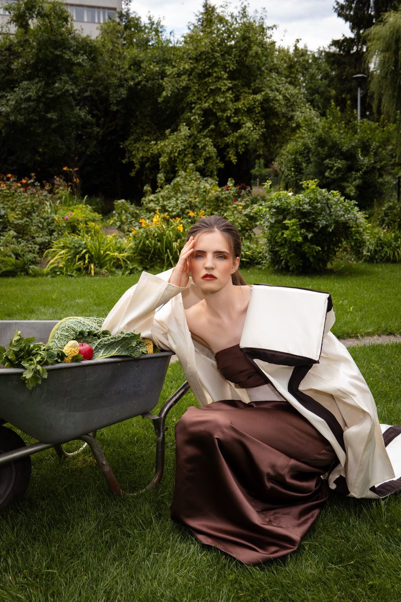 Fashion photo of a woman in an avant-garde dress kneeling beside a wheelbarrow full of vegetables in a green garden landscape.