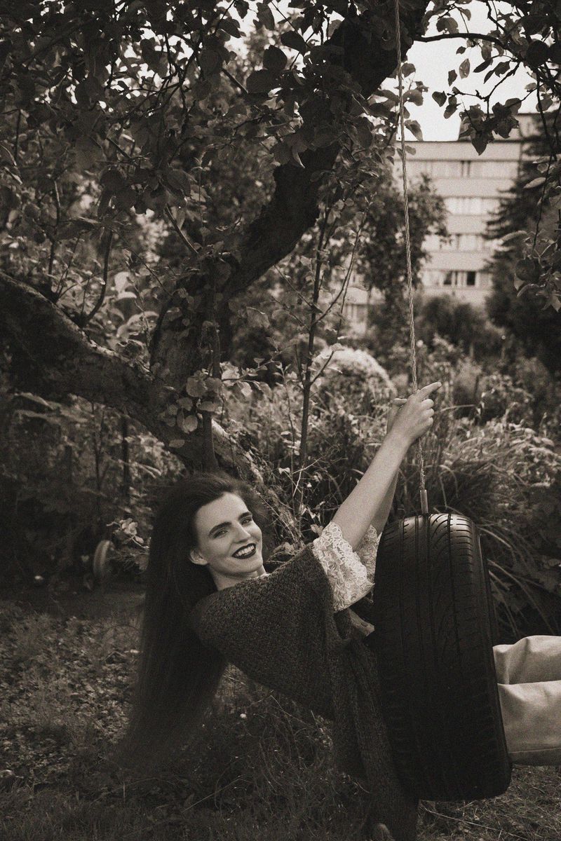 Woman in vintage-style dress on a tire swing under tree branches in sepia tone with building in background, smiling and looking at camera.