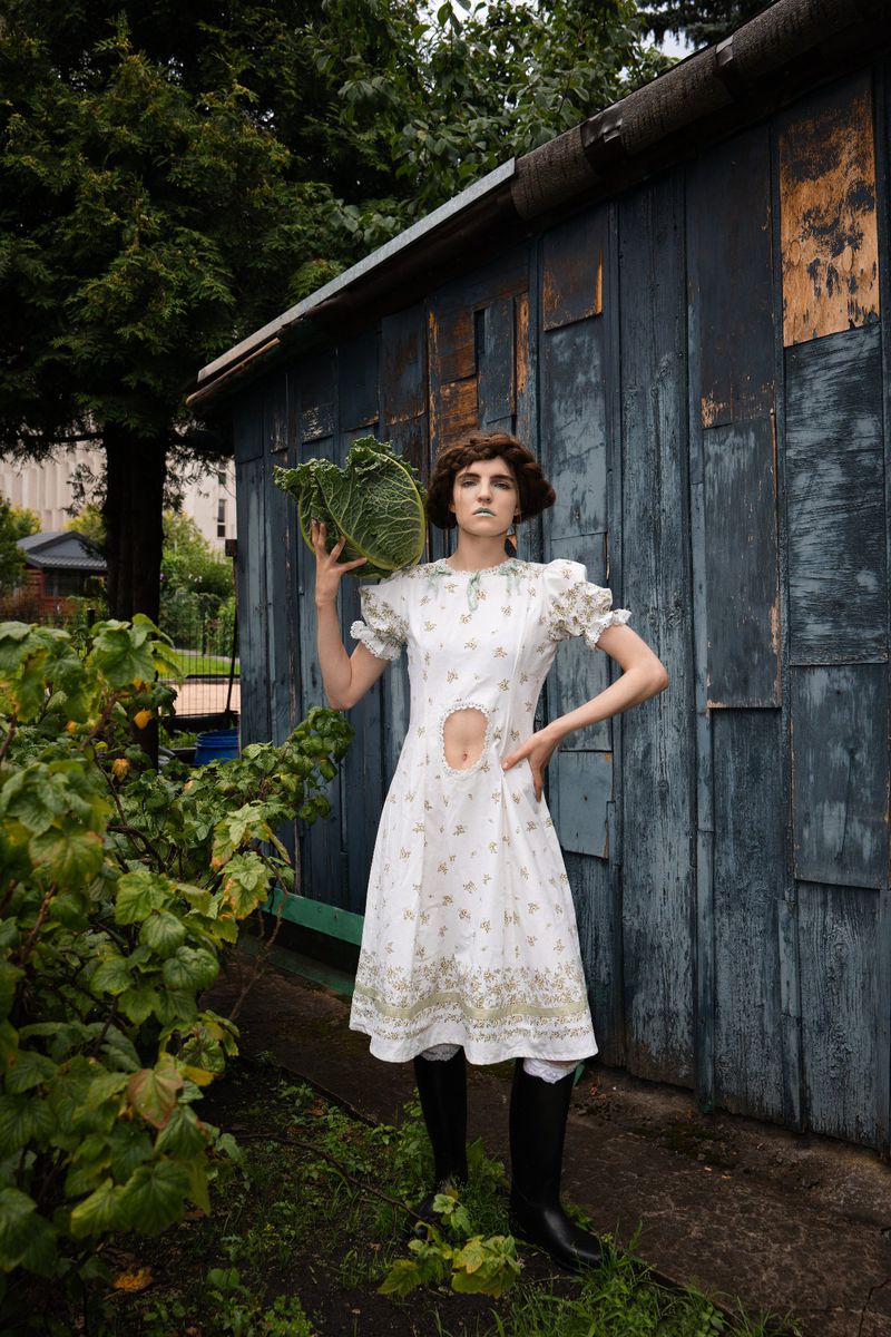 A slender woman in a white floral dress and black boots poses in a garden with a large green cabbage and a rustic blue shed.