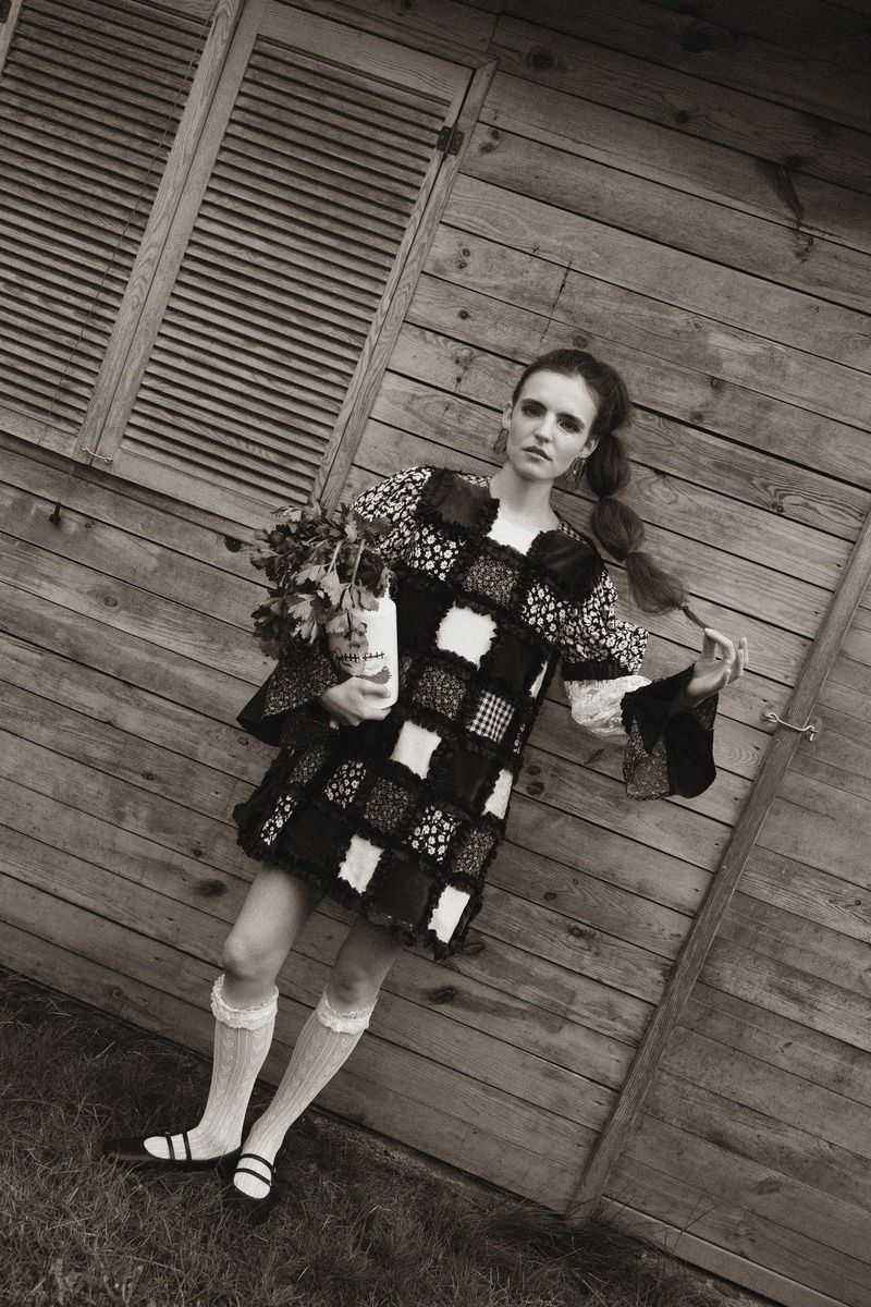 Monochrome portrait of a vintage-styled female model in front of a weathered wood shed, holding flowers with a desaturated color palette.