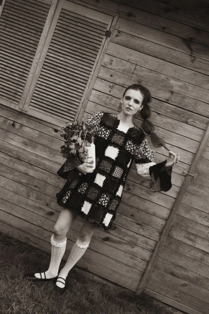 Monochrome portrait of a vintage-styled female model in front of a weathered wood shed, holding flowers with a desaturated color palette.
