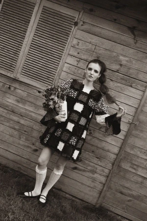 Monochrome portrait of a vintage-styled female model in front of a weathered wood shed, holding flowers with a desaturated color palette.