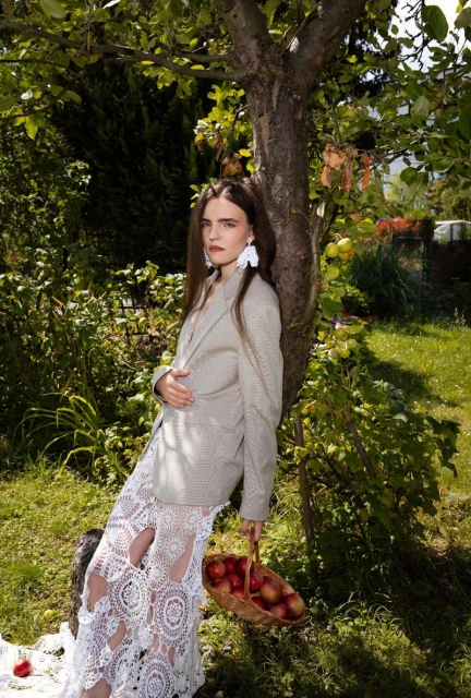 A fashionable young woman stands with a basket of apples under an apple tree in a verdant garden wearing a blazer and lace skirt.