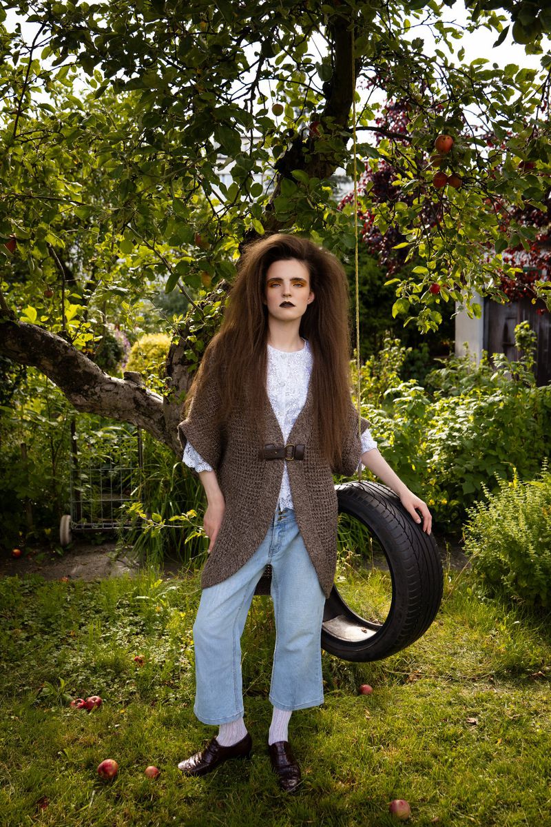 A young woman with long brown hair poses in a garden, wearing a cardigan, jeans, and brown shoes, with a tire swing in the background.
