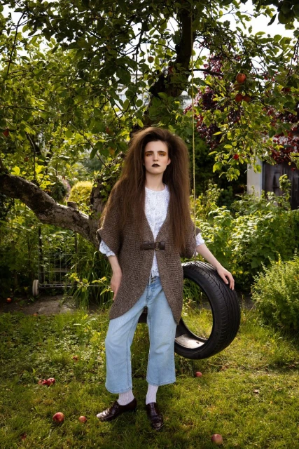 A young woman with long brown hair poses in a garden, wearing a cardigan, jeans, and brown shoes, with a tire swing in the background.