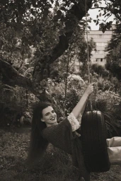 Woman with long dark hair smiling while swinging on a tire swing suspended from a tree branch in a garden
