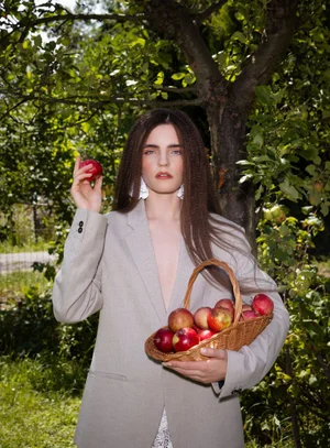 Model in grey blazer holding an apple and a woven basket of apples in a garden