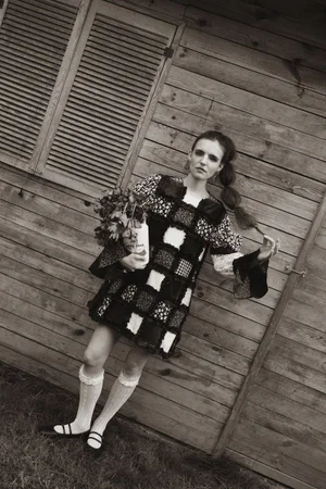 A woman posing in a patchwork mini dress, knee-high socks, and mary janes against a rustic wooden shed wall.