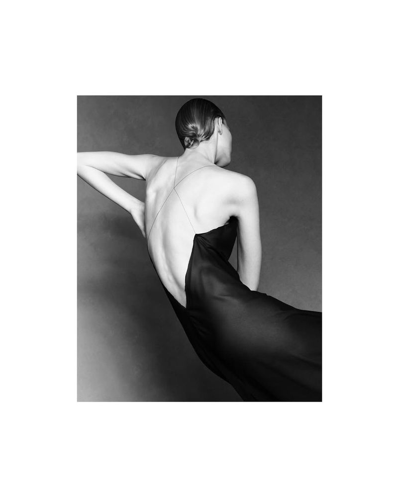 Back view of a woman with styled hair wearing a black evening gown with crossed straps, against a dark grey background.