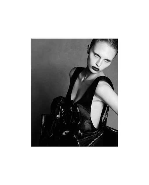 Black and white studio portrait of a woman with slicked-back hair wearing a shiny black top and dark lipstick against a grey backdrop.