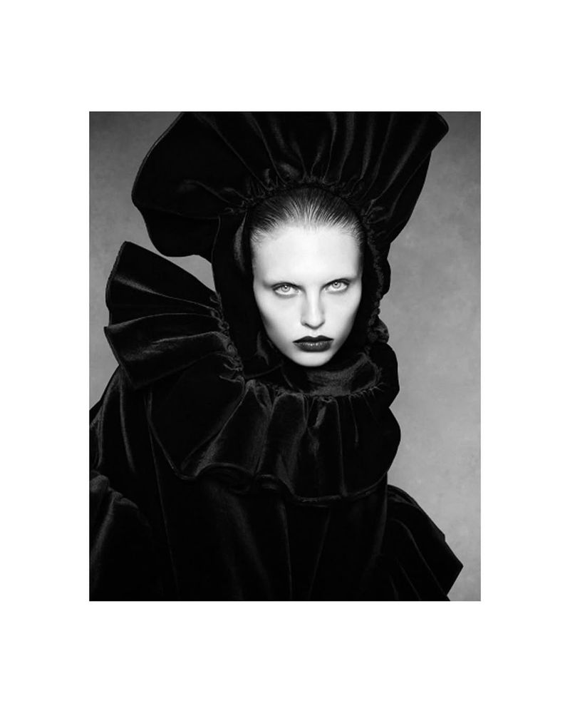 Monochrome portrait of a fair-skinned woman with dark lipstick wearing a large, ruffled black velvet hood and gown against a grey backdrop.