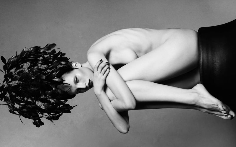 Black and white artistic portrait of a reclining adult woman with feathers in her hair, resting on a black leather surface in studio.
