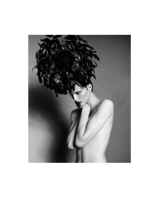 Black and white portrait of a topless fashion model wearing a large feathered headpiece against a grey background in a studio setting.