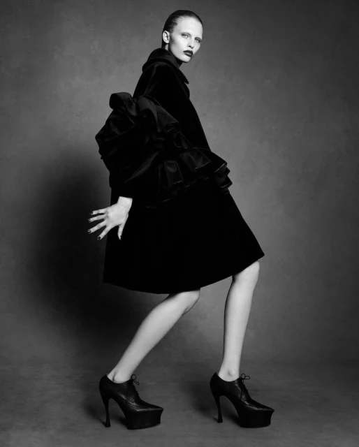 A model in a black coat with frilled sleeves and high heels poses against a grey background in a high-fashion black-and-white photograph.