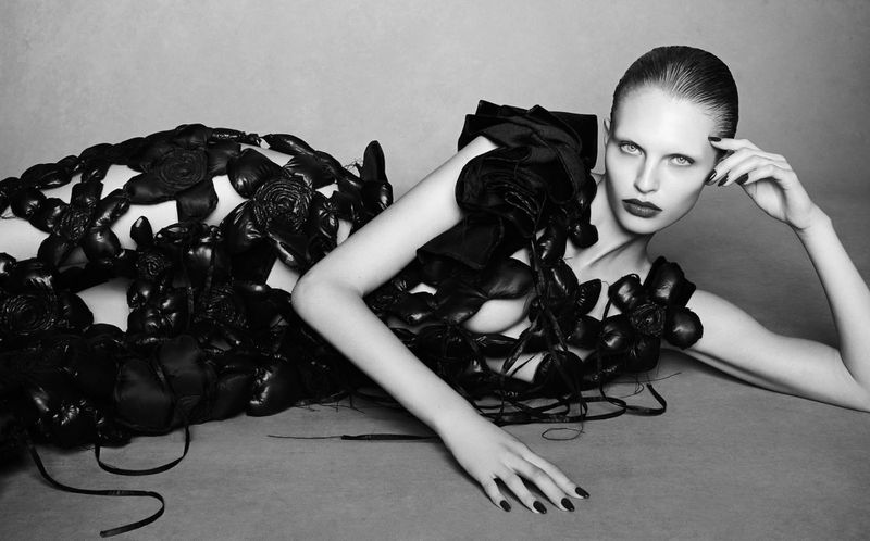Fashion model in a black gown decorated with black floral elements, lying on a grey surface in a studio setting, editorial shot.