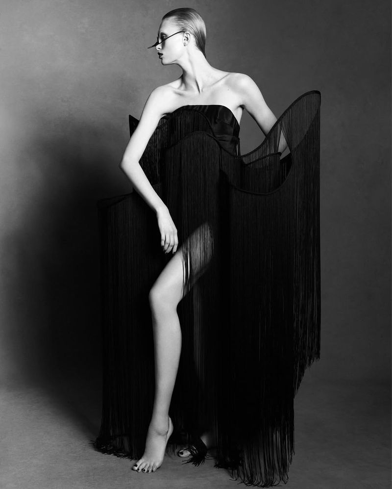 A woman poses in a black strapless dress with fringe, complemented by glasses, set against a gray backdrop in a studio environment.