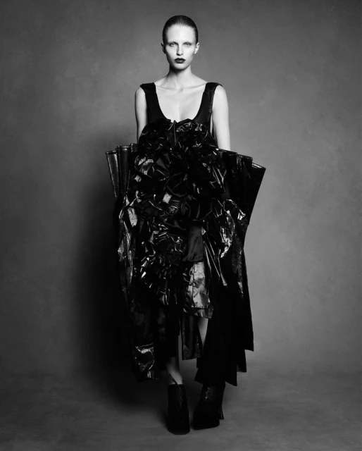 Black and white fashion portrait of a female model in a dramatic dark dress, striking pose against a muted grey background in studio lighting.