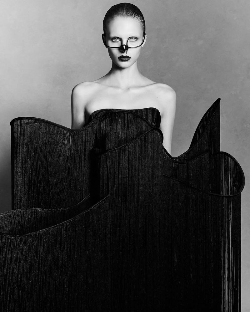 Portrait of a fair-skinned adult female wearing a black, strapless garment and decorative eyewear against a grey background.