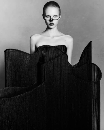 Portrait of a fair-skinned adult female wearing a black, strapless garment and decorative eyewear against a grey background.