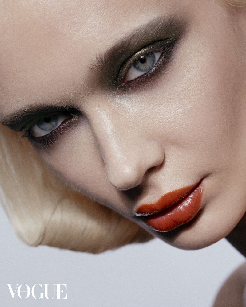 Close-up of a woman's face with dramatic makeup; dark smoky eyeshadow, coral-red glossy lips, short blonde hair, beauty shot, Vogue logo.