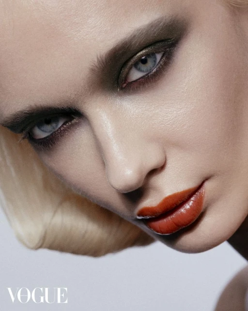Close-up of a woman's face with dramatic makeup; dark smoky eyeshadow, coral-red glossy lips, short blonde hair, beauty shot, Vogue logo.