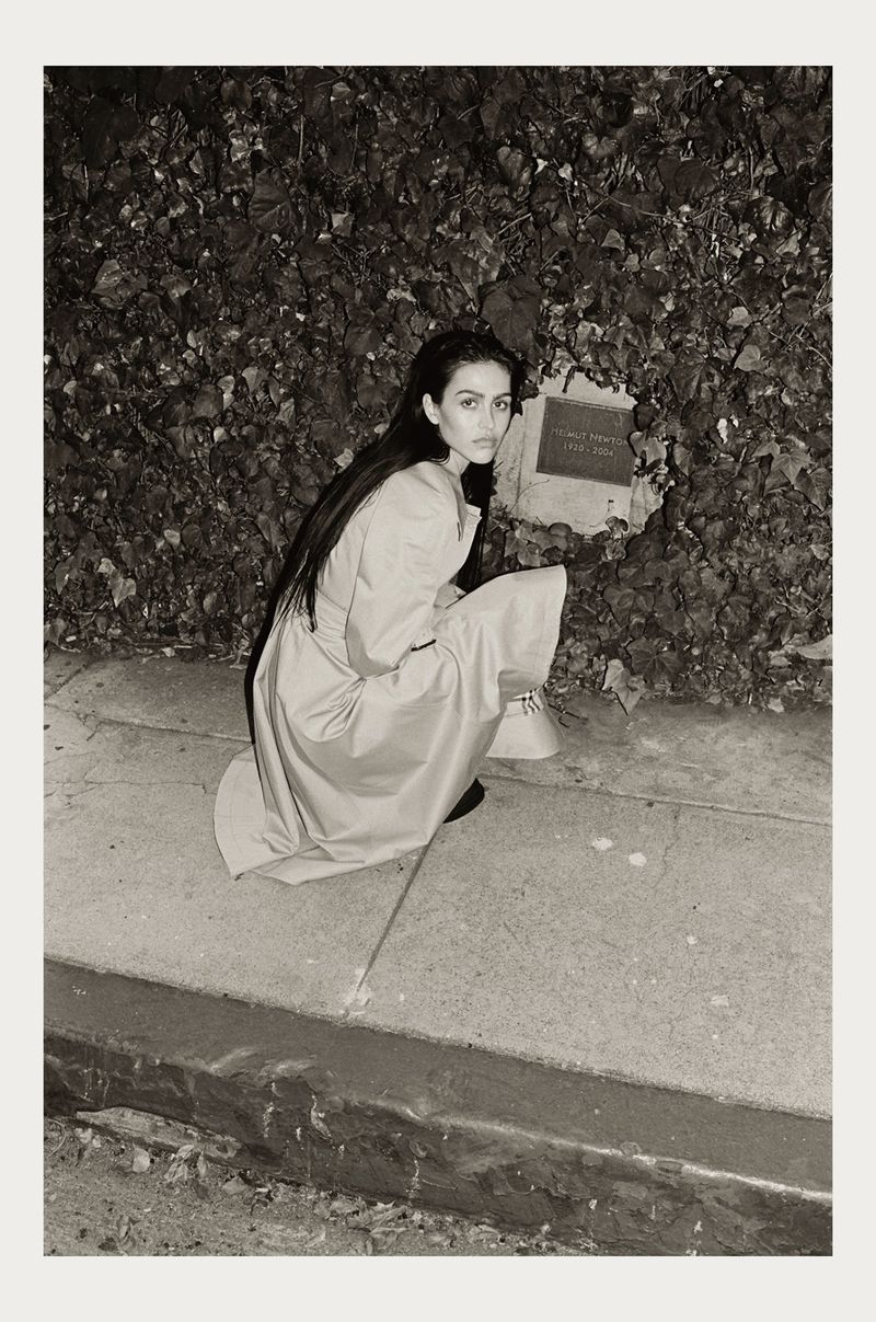 Monochrome portrait of a woman in a light dress crouching on a sidewalk next to a plant-covered wall, looking directly at the camera.