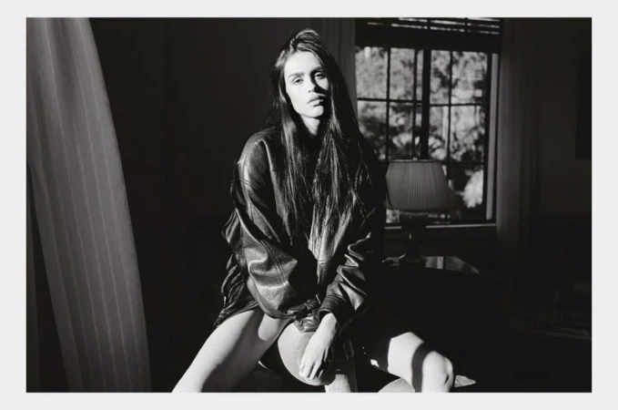 Portrait of a young woman with long brown hair wearing a black leather jacket sitting in a room with a window and a lamp.