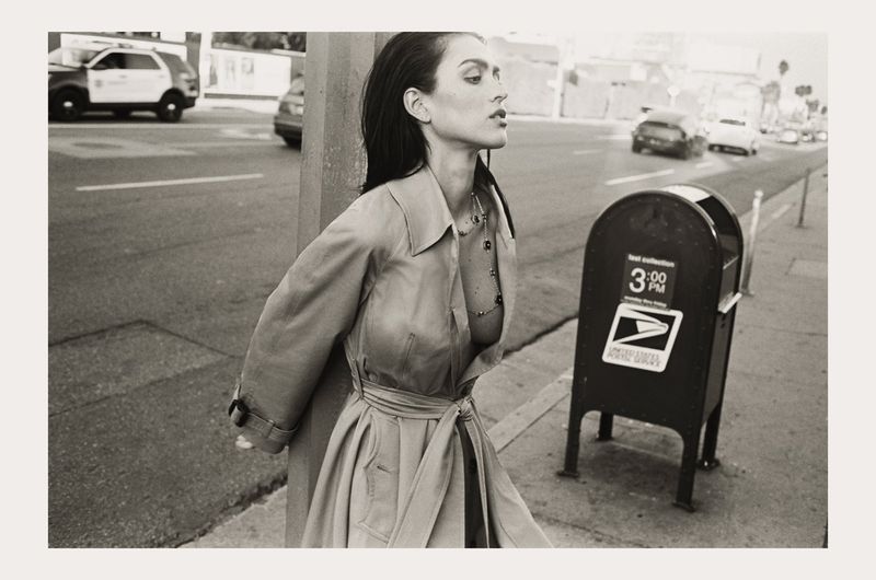 Monochrome portrait of a woman in a trench coat standing on a city sidewalk with a mailbox, vehicles, and buildings in the background.