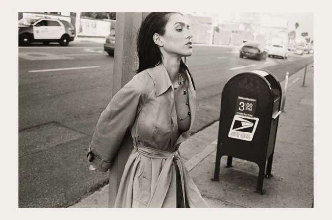 Monochrome portrait of a woman in a trench coat standing on a city sidewalk with a mailbox, vehicles, and buildings in the background.