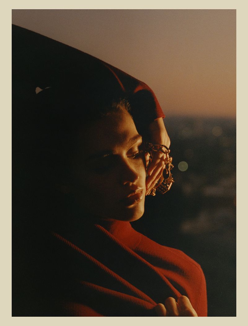 Adult woman wearing red turtleneck sweater and jewelry poses in front of city skyline, gazing off-camera at golden hour.