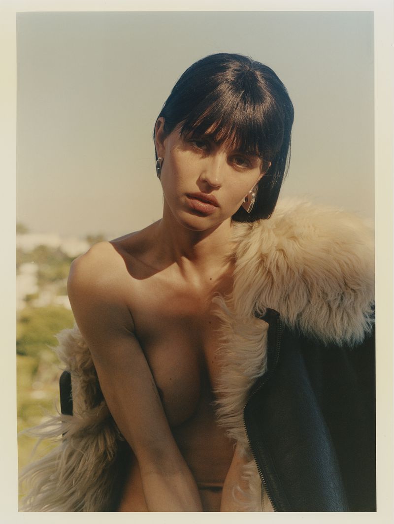 Attractive adult woman with short brunette hair poses in a fur-lined coat in front of an outdoor background under bright lighting.