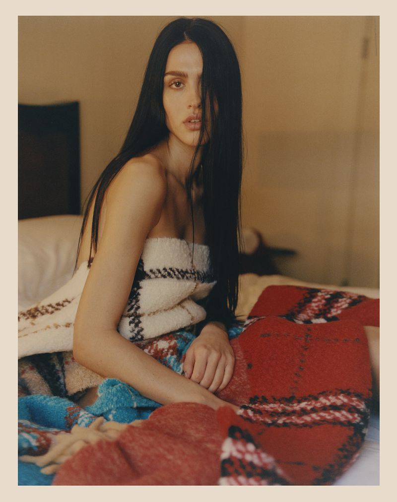 Portrait of a young woman with long dark hair wearing a woven blanket, seated on a bed with additional blankets in a warm indoor setting.