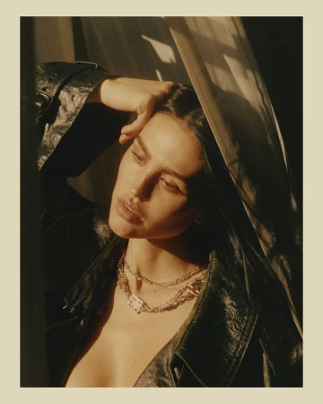 Indoor portrait of a young adult woman with dark hair, wearing a black leather jacket and layered necklaces, with window light.