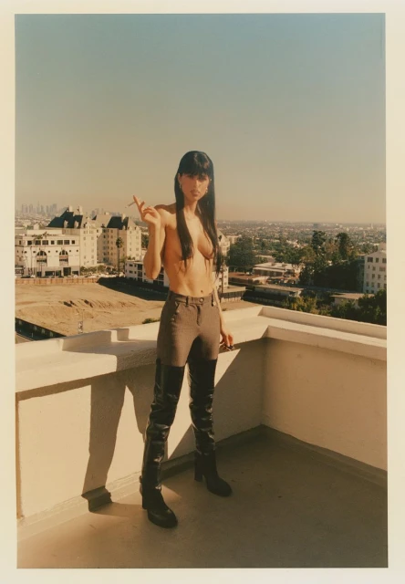 A slender, light-skinned woman stands on a balcony, wearing a top, pants, and thigh-high boots, while holding a cigarette outdoors.